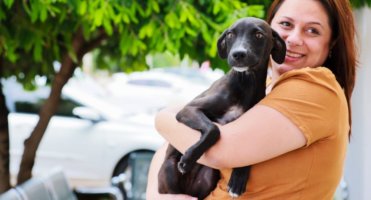 Mulher segurando um cachorrinho no colo e sorrindo para a câmera. Árvore ao fundo e carro estacionado.