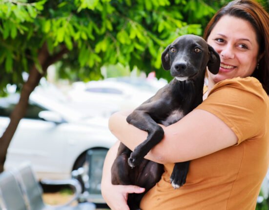 Mulher segurando um cachorrinho no colo e sorrindo para a câmera. Árvore ao fundo e carro estacionado.