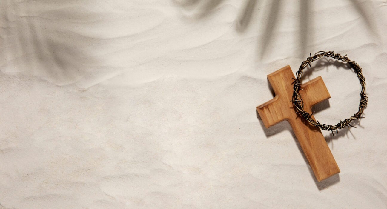 Cruz de madeira em miniatura com cruz de espinhos sobre. Fundo aparenta ser um tecido bege.