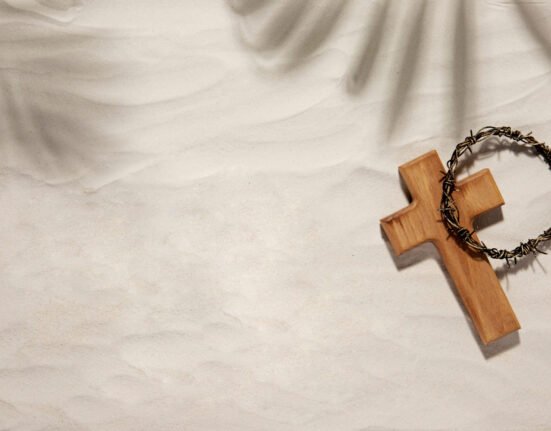 Cruz de madeira em miniatura com cruz de espinhos sobre. Fundo aparenta ser um tecido bege.